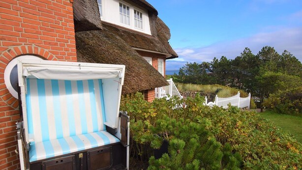 A thatched-roof house with a white picket fence, a striped outdoor seating area, and a well-maintained garden.