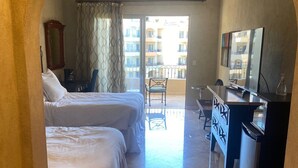 Habitación de hotel con dos camas, un escritorio, una silla, un televisor y un balcón con vistas a edificios.