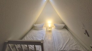 Un dormitorio abuhardillado pequeño y triangular con dos camas individuales, una mesita de noche con lámpara y una silla de madera.