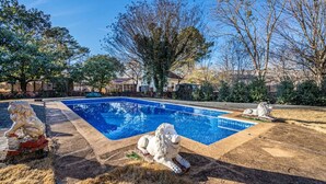 Una piscina rectangular con un borde de baldosas, rodeada de hierba seca y dos estatuas de leones de piedra.