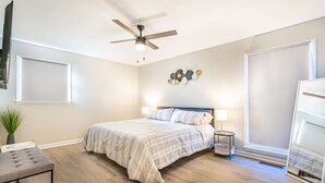 Un dormitorio con cama, ventilador de techo, mesita de noche con lámpara, planta en maceta y ventana con persianas.