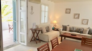 Een woonkamer met een bank, een salontafel en een eettafel. Een deur leidt naar een buitenruimte met een stoel.