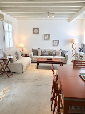 Un salon comprenant un canapé, une table basse et une table à manger. Il y a des lampes fixées au mur et une fenêtre avec des rideaux.