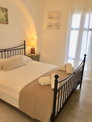 Un lit bien fait, avec du linge de lit blanc et un plaid beige, une lampe de chevet et une table de chevet avec une autre lampe.
