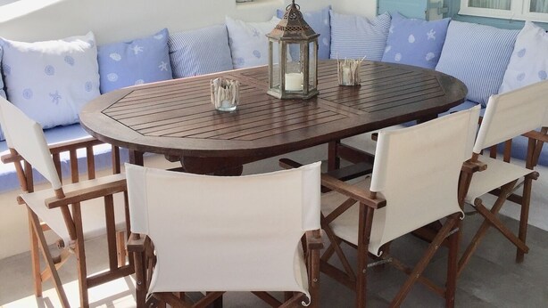 Een ronde houten tafel met witte stoelen met kussens, een hanglamp en een blauwe deur op de achtergrond.