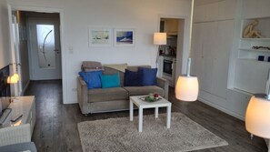 Un salon avec un canapé, une table basse et une lampe. On aperçoit une cuisine au fond.