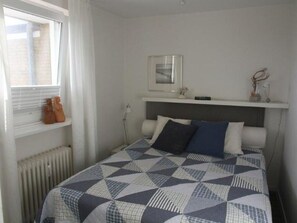 Une chambre à coucher avec un lit, une fenêtre, un radiateur, une étagère avec des objets et un tableau encadré au mur.
