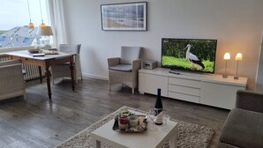 Un salon comprenant un téléviseur, une table à manger, des chaises, un canapé et une table basse sur laquelle se trouvent une bouteille et des verres.