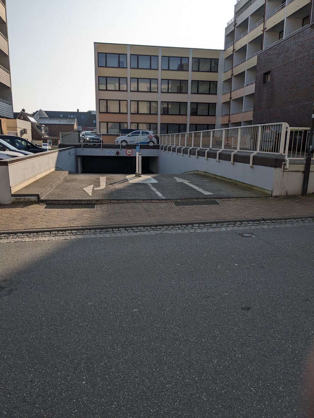 Un espace de stationnement doté d’une entrée fermée par un portail, une voiture garée à l’intérieur, et un bâtiment à plusieurs étages en arrière-plan.