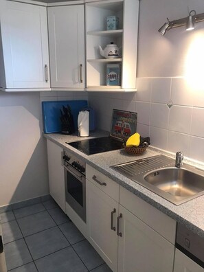 Cozinha compacta com armários brancos, pia de aço inoxidável, fogão e bancada com tábua de corte azul e um cesto de limões.