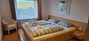 Un letto matrimoniale con biancheria a righe, una poltrona in vimini, un comodino con una lampada e una finestra con tende.