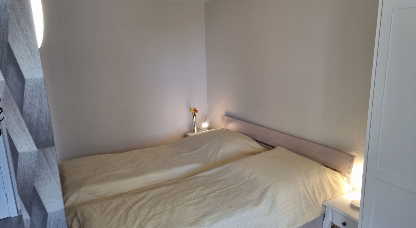 Un dormitorio sencillo y sin adornos, con cama, mesita de noche y velador con lámpara.