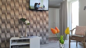 A modern living room with a geometric-patterned wall, a flat-screen TV mounted above a white console, a vase with flowers, and a wicker chair with a cushion.