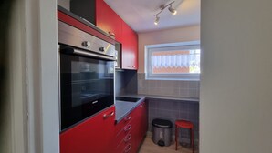 A compact kitchen with red cabinets, a Siemens oven, a window with lace curtains, and a small stool.