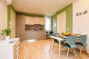 A modern kitchen with wooden cabinets, a dining area with green chairs, and a breakfast setup with fruit and coffee. - Appartamento Ulivo (Salò)