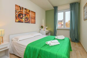 A hotel room with a bed, a nightstand, a lamp, a window with curtains, and artwork on the wall. - Appartamento Ulivo (Salò)