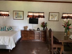 A hotel room with a bed, a wooden dresser, a television, a dining table with chairs, and artwork on the walls.