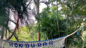 A hammock with the text "HONDUKAR" in blue letters, set against a backdrop of lush greenery and palm trees.