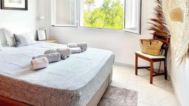 A bedroom with a bed, a window with a view of greenery, a chair, and a basket on a side table. - Beach house 2 Palmeras with pool in Port Pollensa (Port de Pollença)