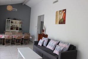 A living room with a grey sofa, a white coffee table, and a dining area with a table set for four. - Single-storey villa, large plot, close to lake and ocean (Hourtin)