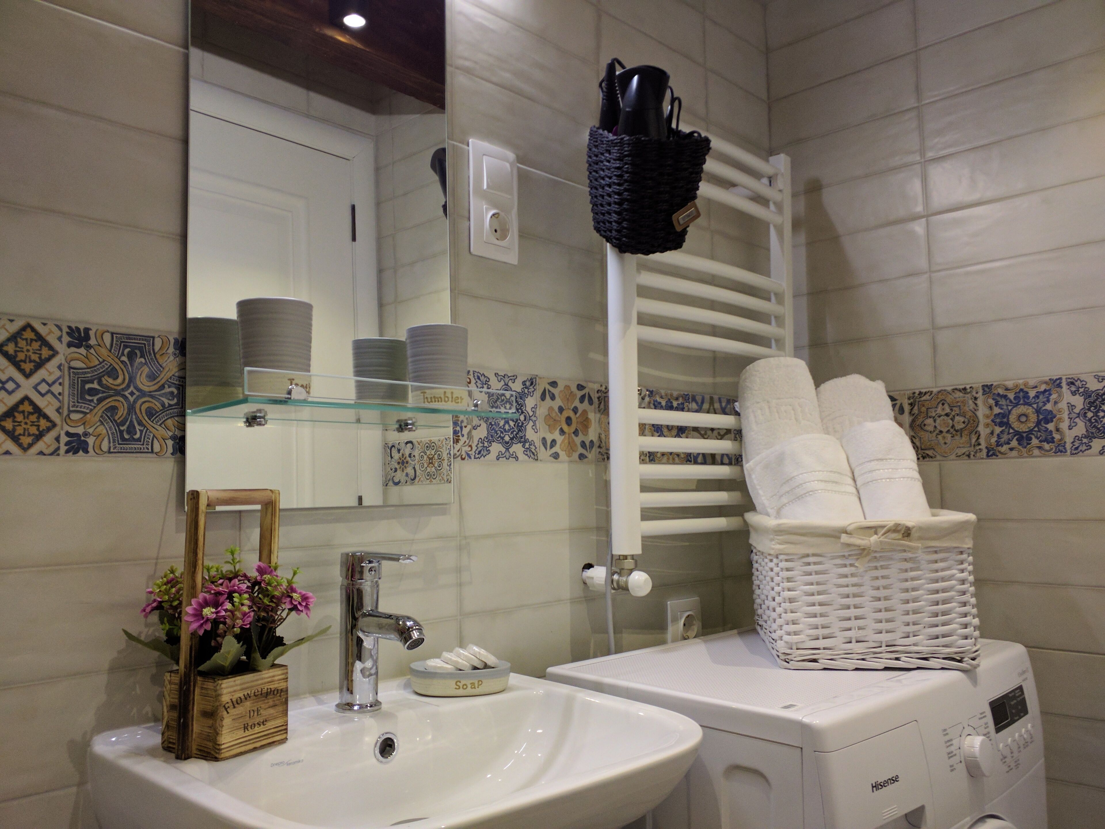 Un baño con un lavabo blanco, un espejo, una estantería de vidrio con artículos de tocador, un perchero blanco y una lavadora Hisense.