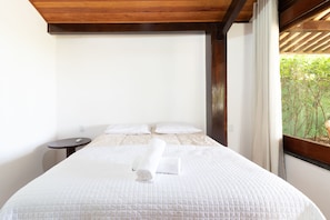 A neatly made bed with white bedding and pillows, a small bedside table with a remote control, and a window with a view of greenery. - GB51 Excelente Casa 4 Suítes Vista Mar (Camaçari)