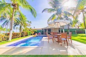 A swimming pool with a tiled deck, surrounded by palm trees and a patio area with wooden chairs and a table. - GB51 Excelente Casa 4 Suítes Vista Mar (Camaçari)
