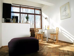 Ein Zimmer mit großem Fenster, Schreibtisch, Stuhl und einem dunkel gefärbten Pouf.