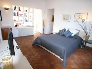 Ein Schlafzimmer mit einem großen Bett, einem Schreibtisch mit Fernseher, einem Heizkörper und einem Regal mit diversen Gegenständen.