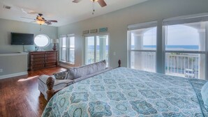 Een slaapkamer met een groot bed, een plafondventilator, een dressoir, een televisie en een balkon met uitzicht op de oceaan.