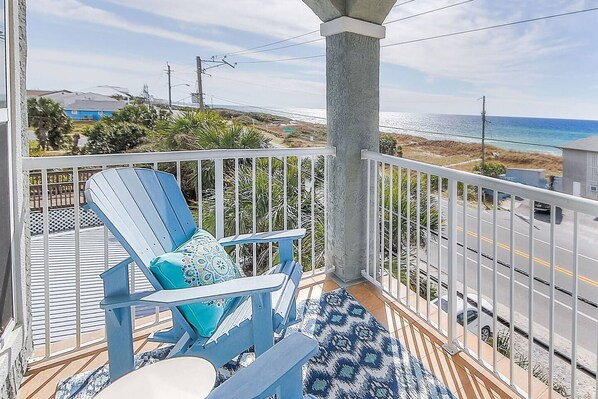Un balcon avec un fauteuil Adirondack bleu, un tapis à motifs et une vue sur l’océan et la plage.
