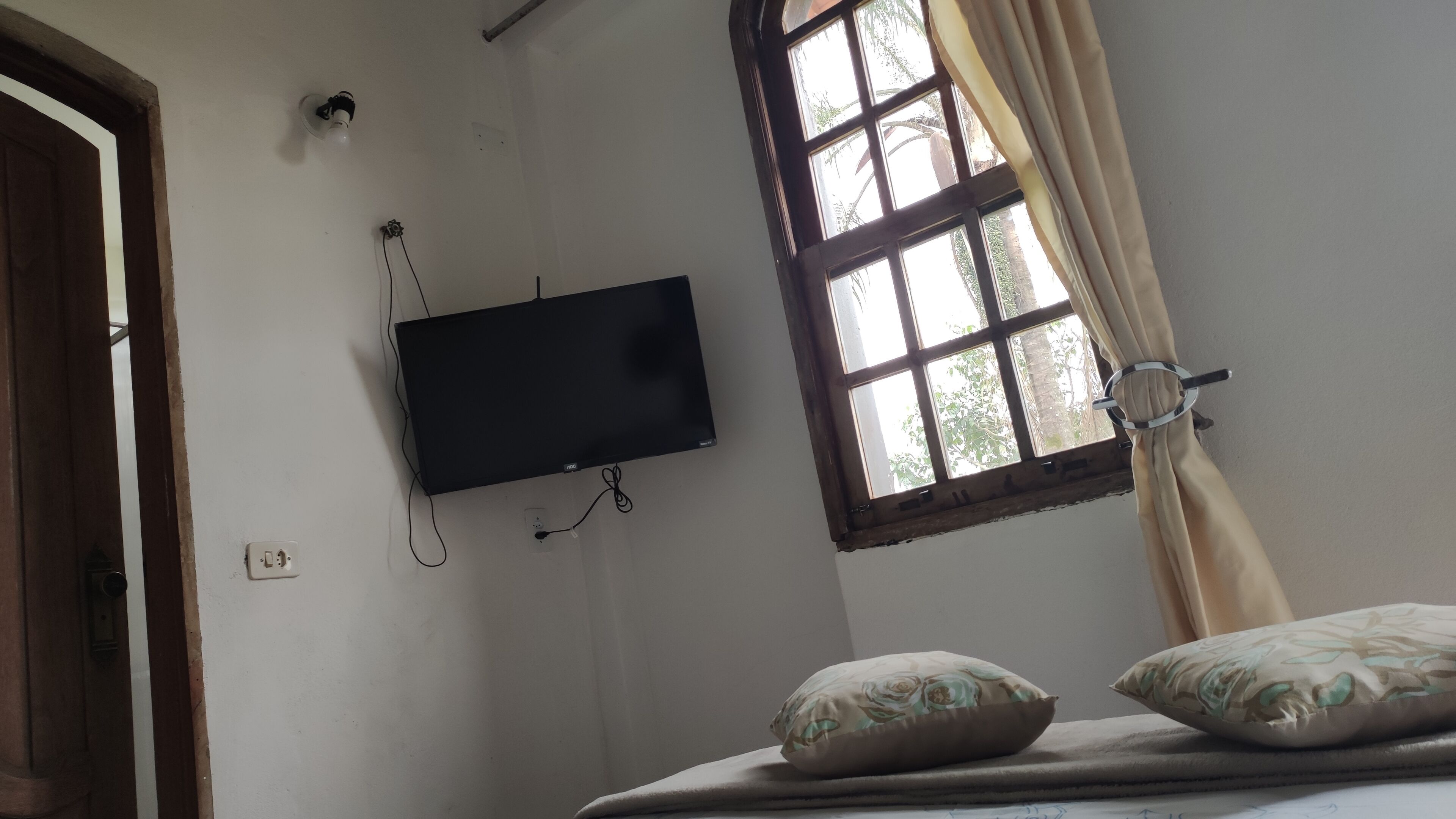 Una habitación con televisión instalada en la pared, una ventana con cortinas y una cama con almohadas.