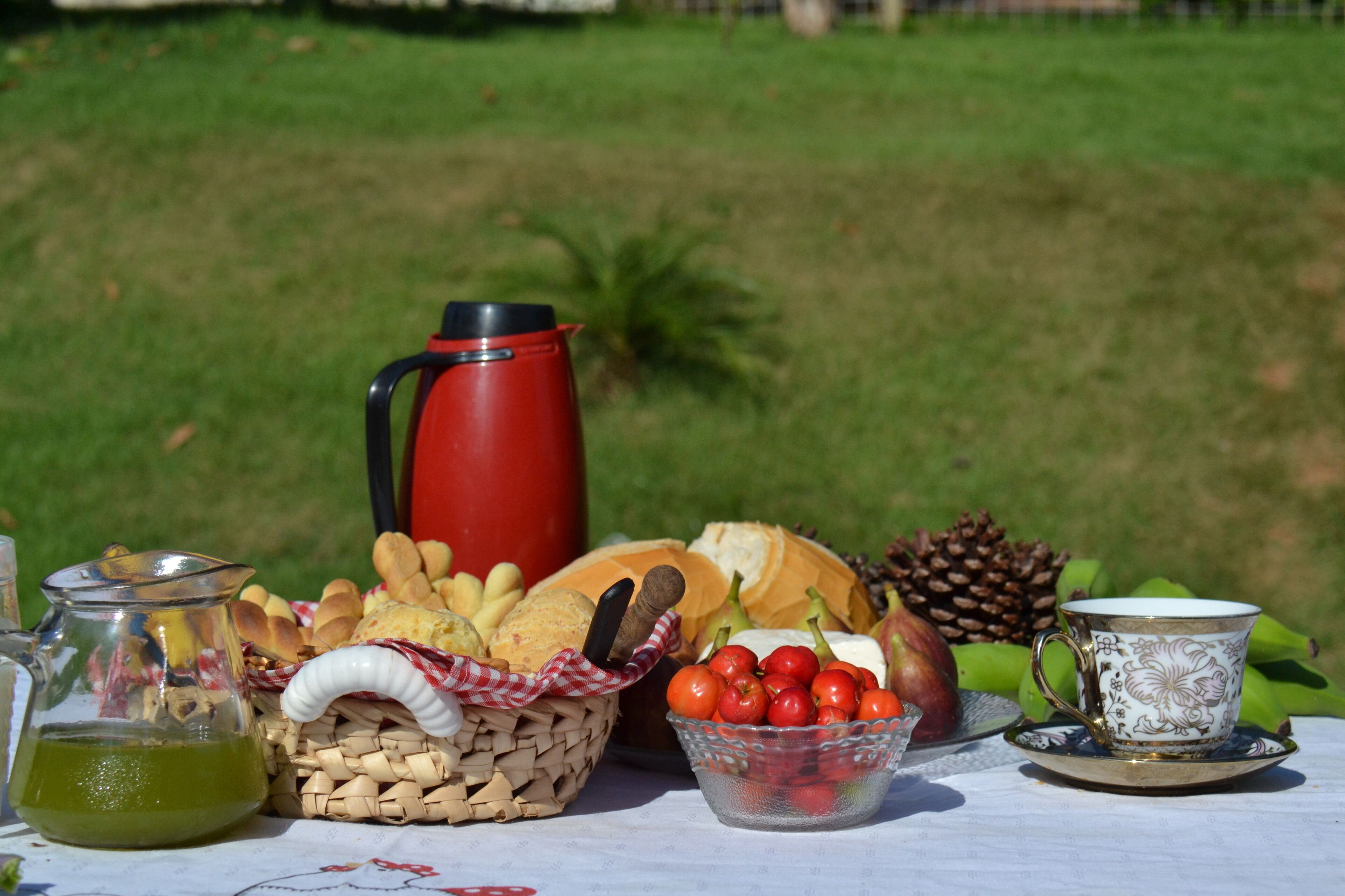 A picnic setup with a red coffee pot, bread, cheese, tomatoes, and a decorative cup on a table.