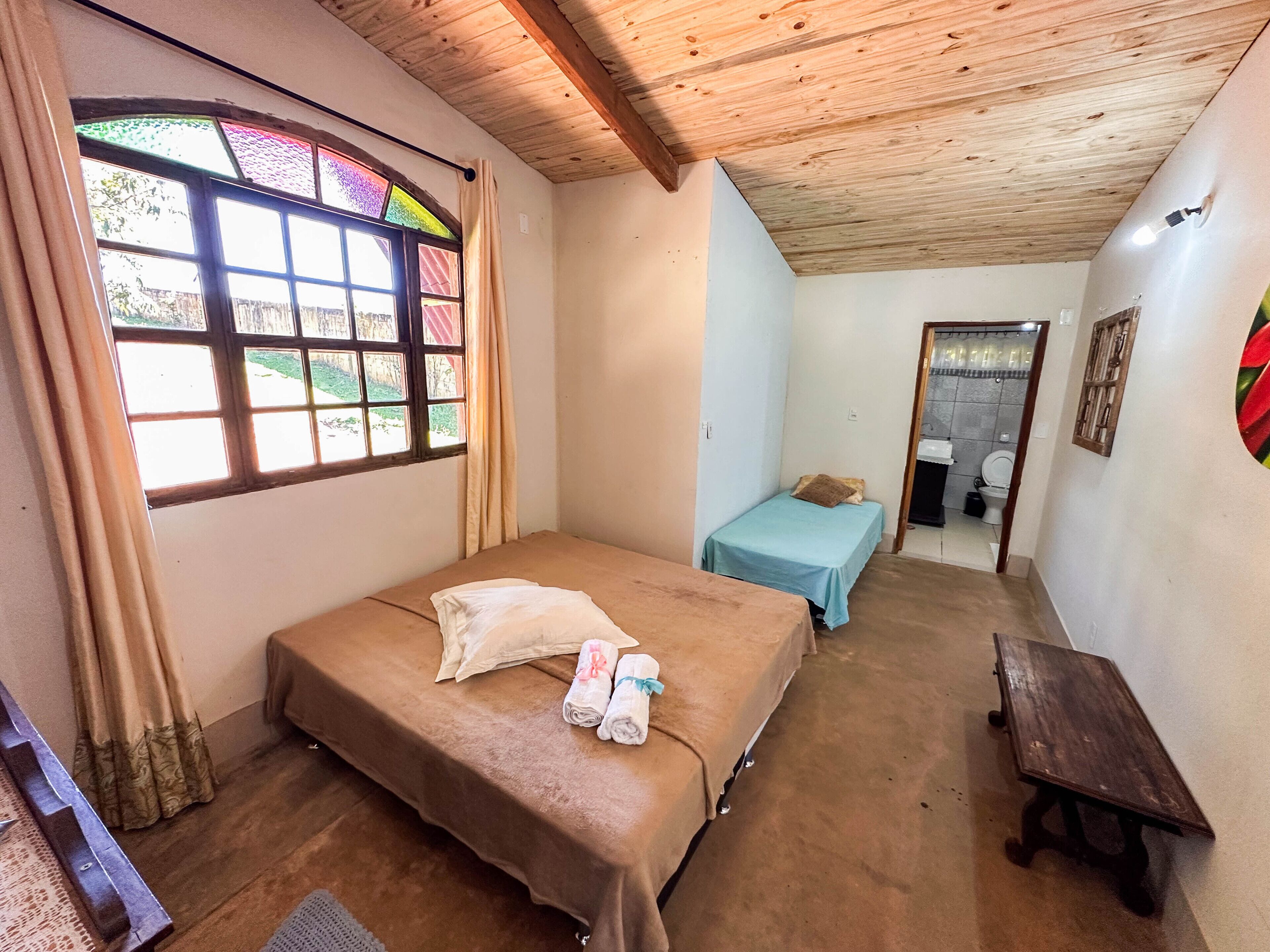 Um quarto com teto de madeira, cama com colcha marrom, uma mesinha e uma janela com cortinas.