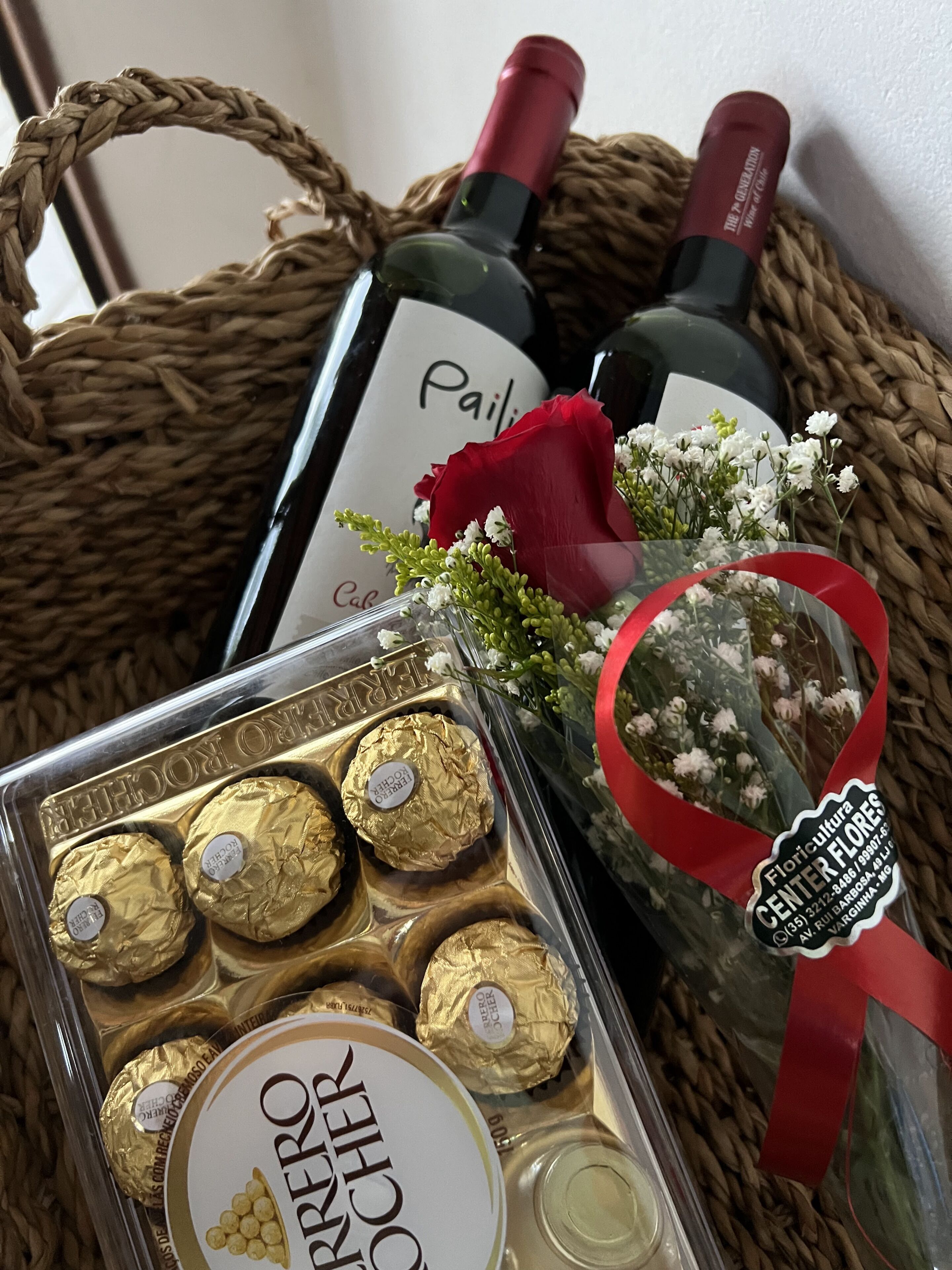 Un cesto in vimini con una bottiglia di vino, una scatola di cioccolatini Ferrero Rocher e un mazzo di fiori.