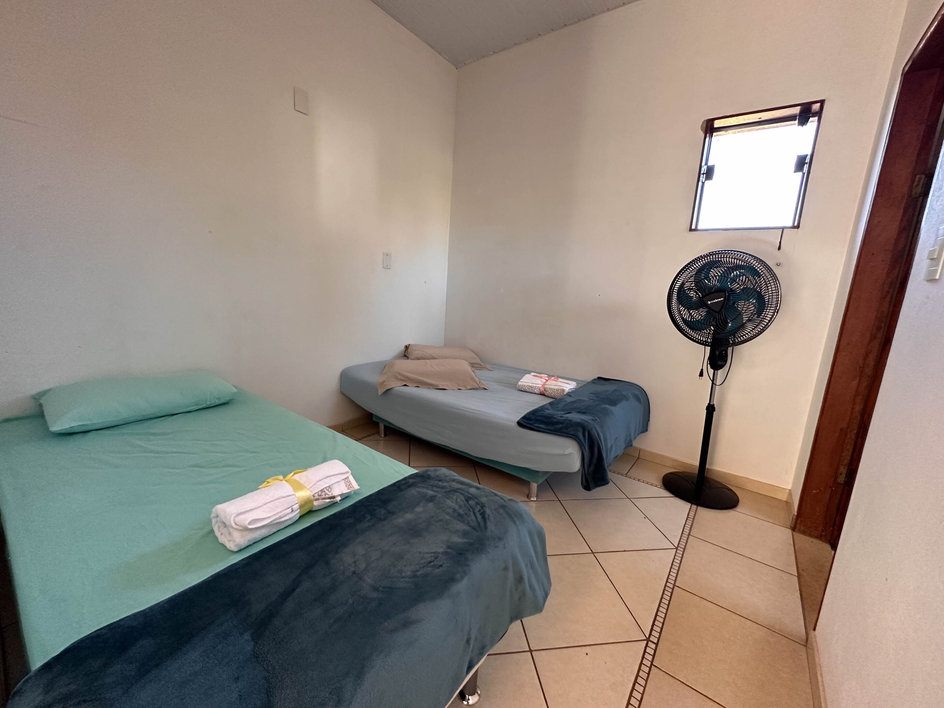 A room with two beds, one with a green bedspread and the other with a blue bedspread, a standing fan, and a small window.