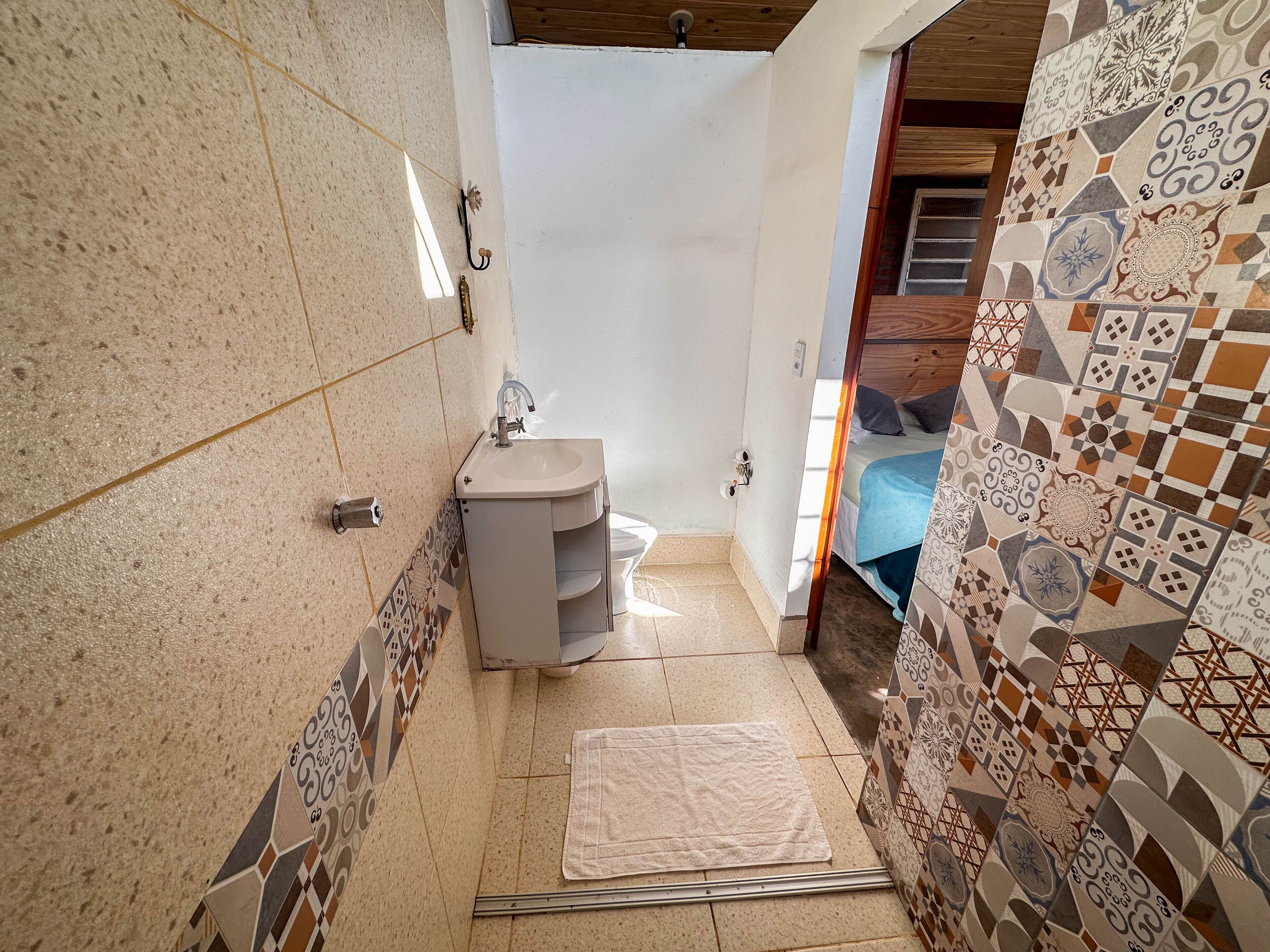 A small bathroom with a sink, a toilet, and a shower area. The walls are tiled with a geometric pattern, and there is a wooden ceiling.