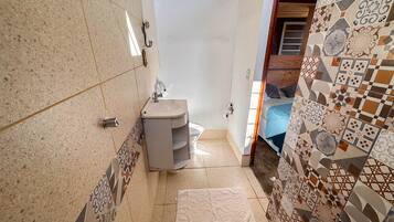 A small bathroom with a sink, a toilet, and a shower area. The walls are tiled with a geometric pattern, and there is a wooden ceiling.
