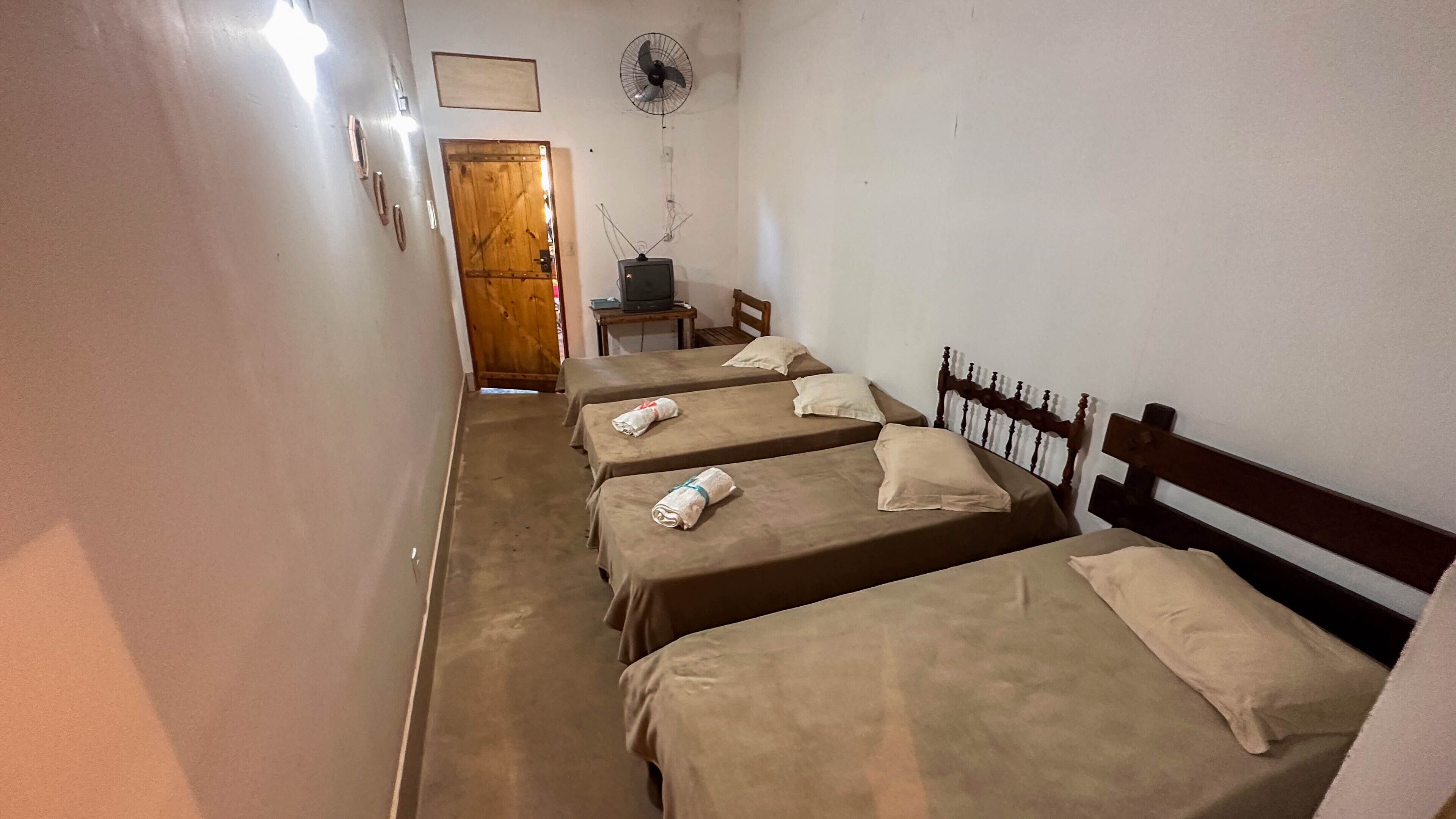 A narrow hallway with four single beds aligned against the wall, each with a wooden headboard and a pillow. A wooden door is visible at the end of the hallway, and a small table with a television is placed against the wall.