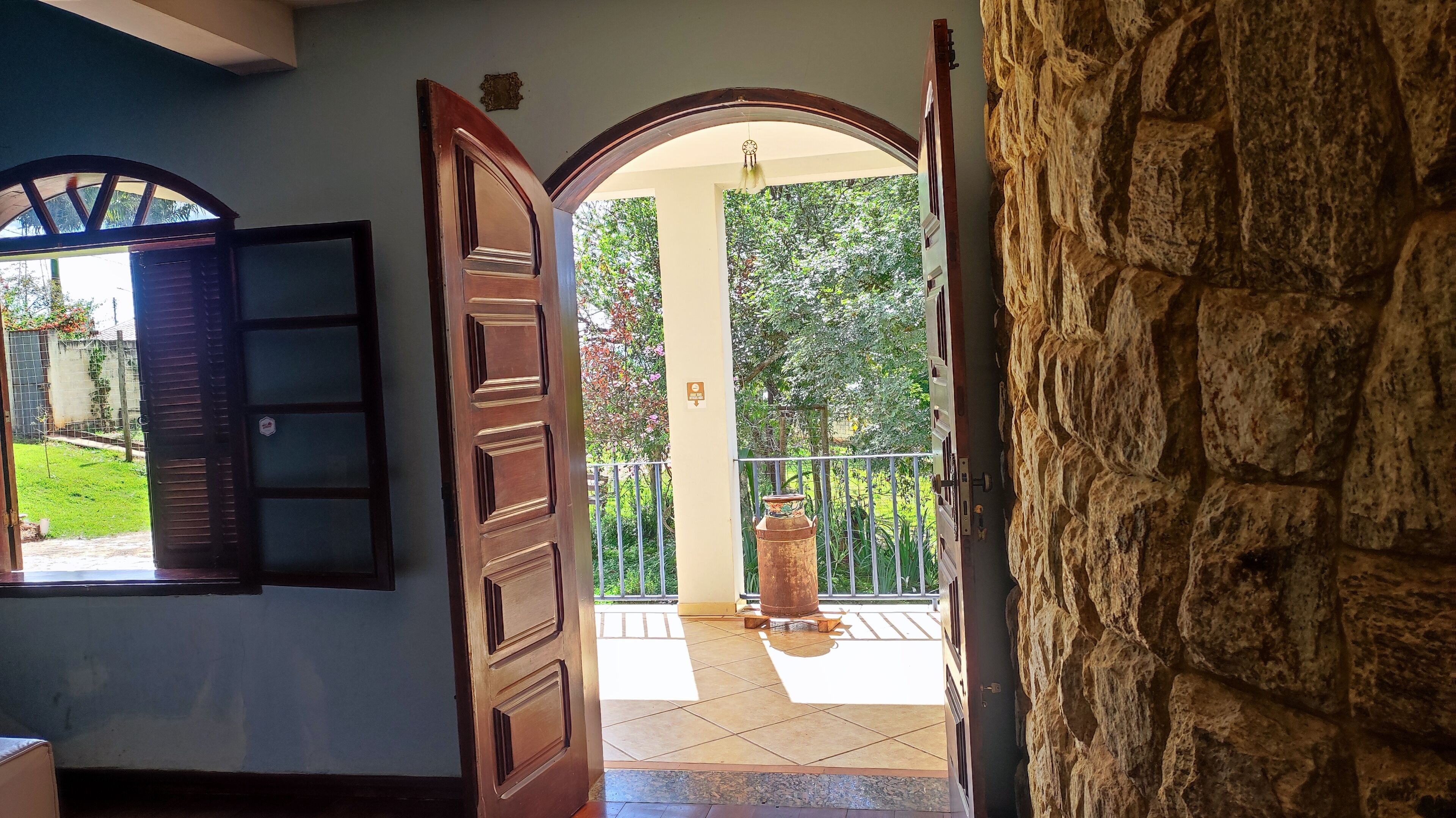 A doorway leading to a balcony with a stone wall, a large wooden door, and a view of a green garden.