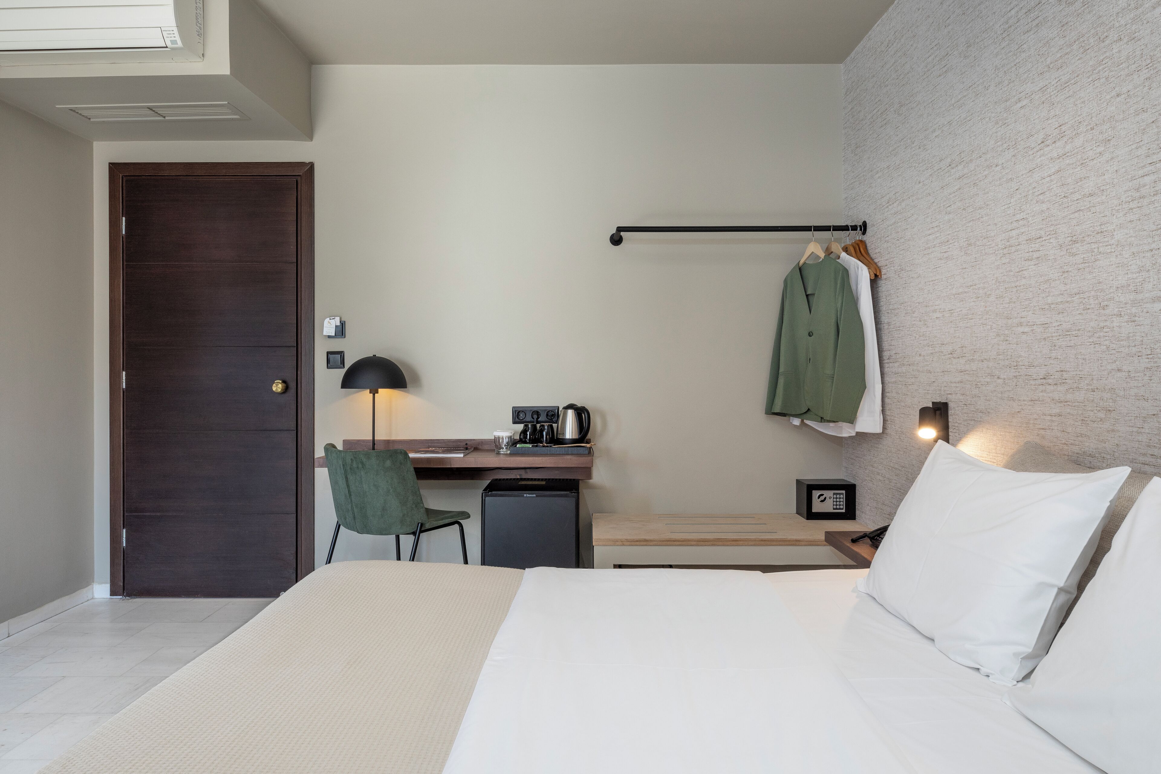 A hotel room with a bed, a desk with a chair, a small refrigerator, a hanging coat rack, and a wall-mounted light.