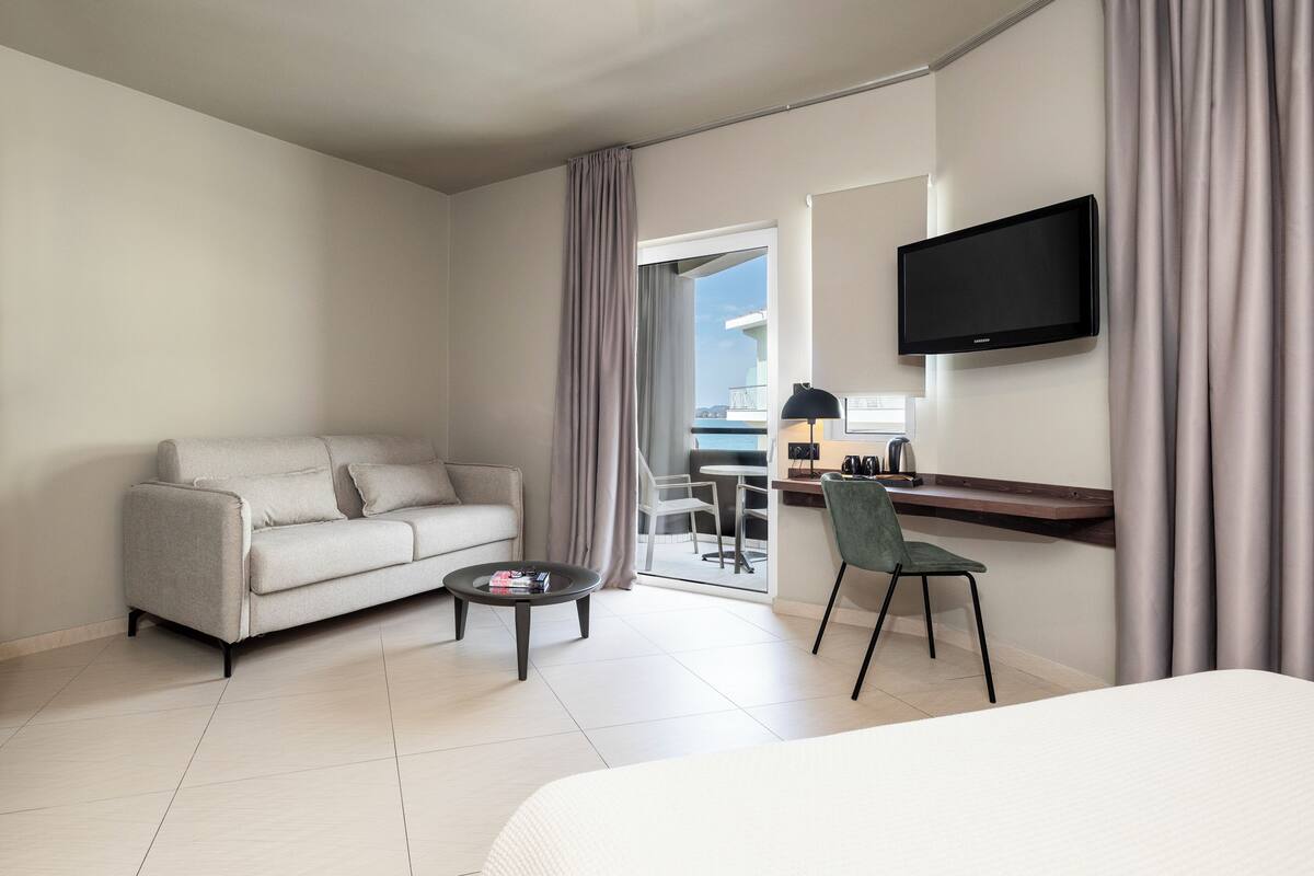 A modern hotel room with a sofa, a small table, a TV mounted on the wall, and a view of the outside balcony.