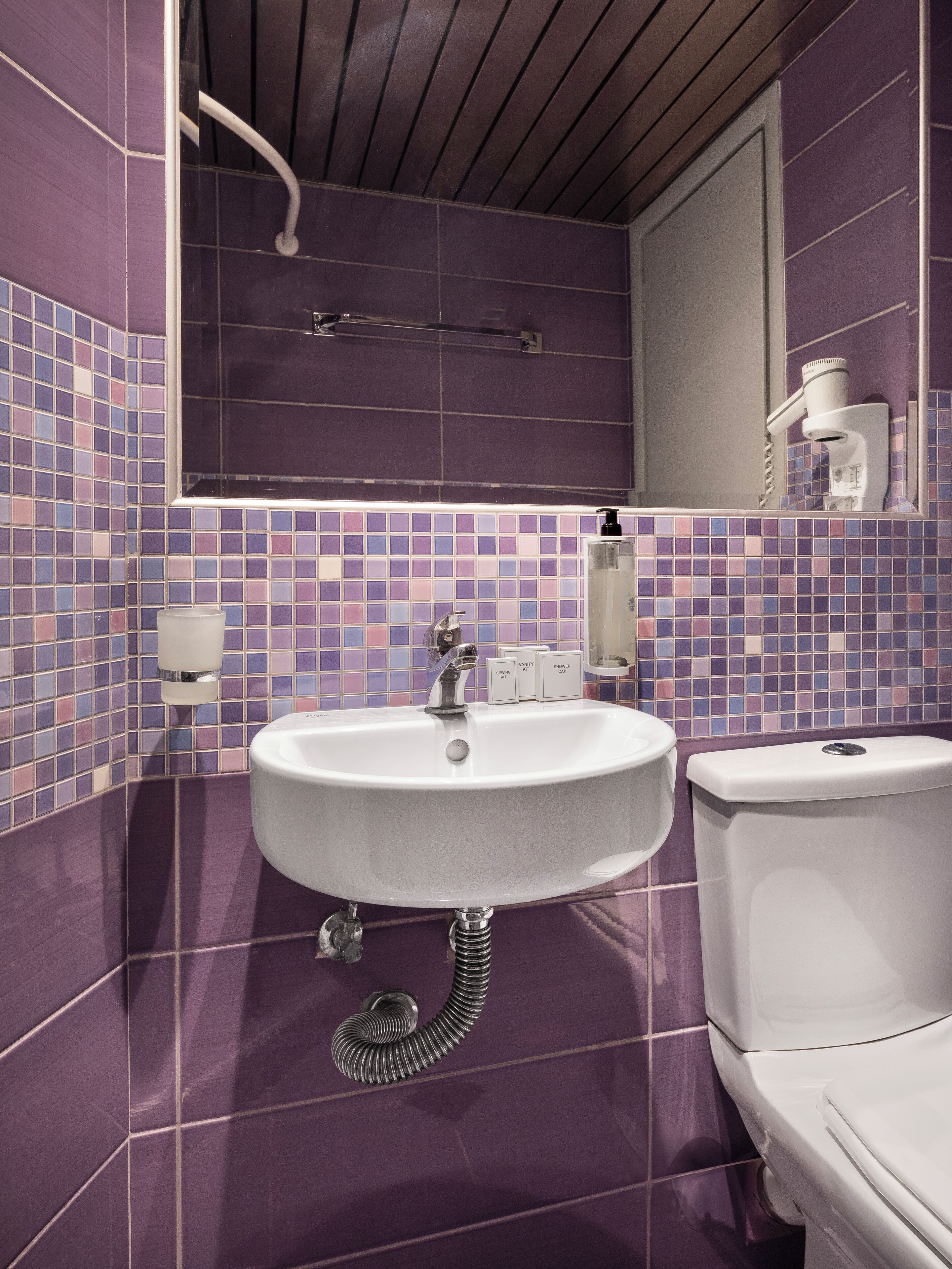 A bathroom with a white sink, a mirror, and a toilet. The walls are tiled with purple and blue tiles.