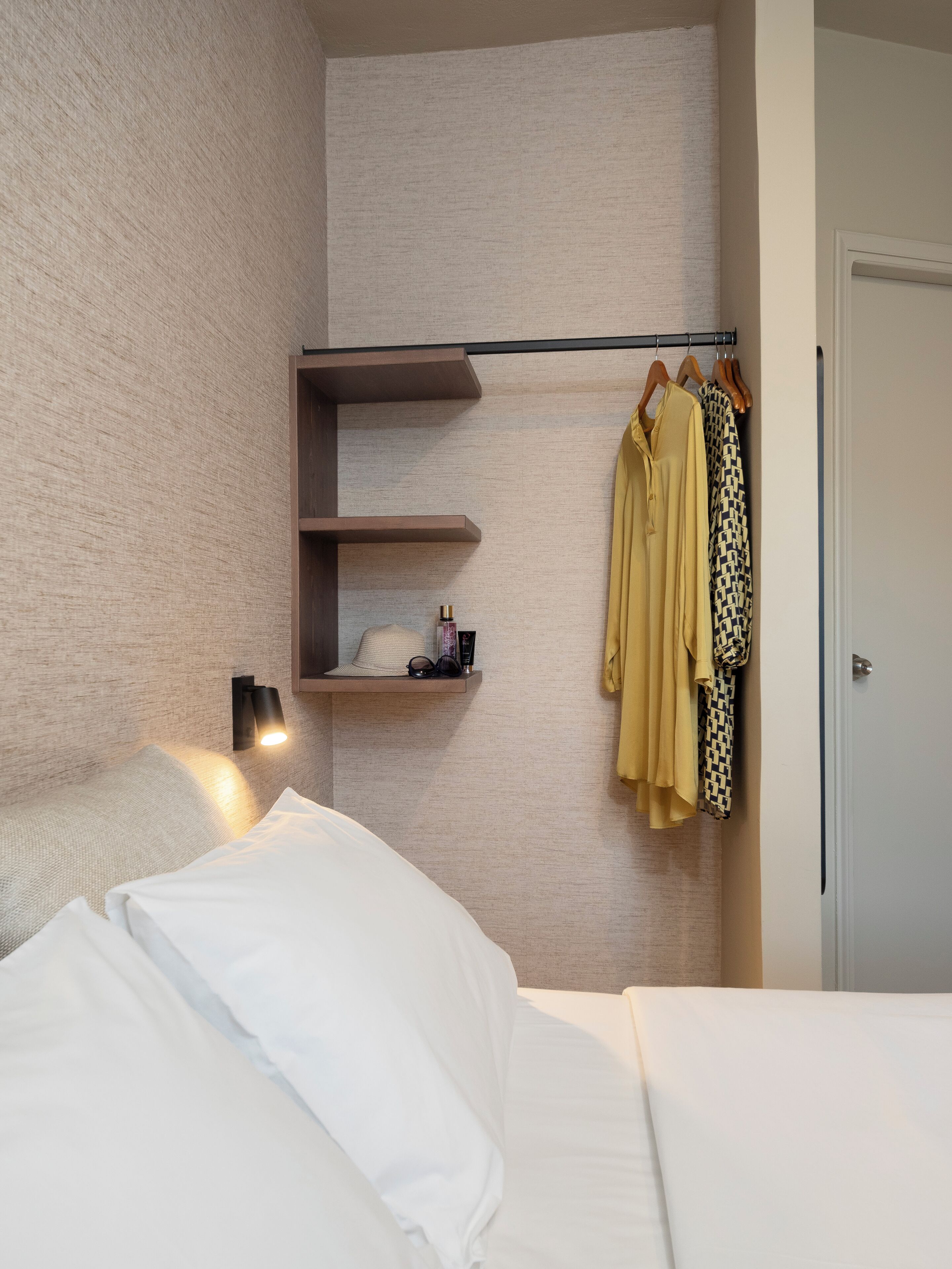 Habitación de hotel moderna con una cama, un estante con un sombrero y gafas de sol, y un perchero con una prenda amarilla.