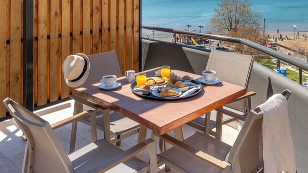 A balcony with a wooden table set for breakfast, overlooking a beach and the sea.