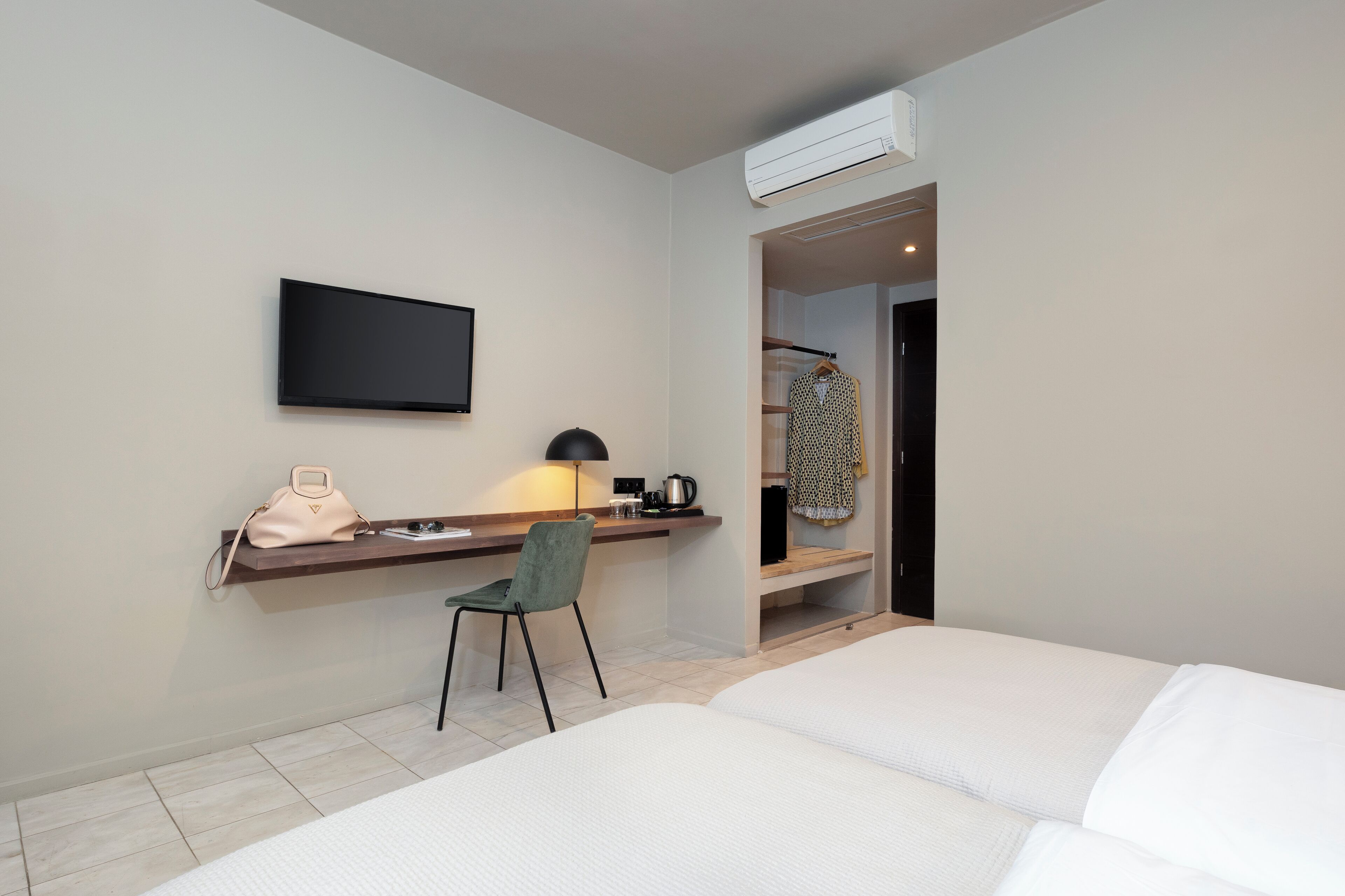 A hotel room with a bed, a desk with a chair, a TV, a coat rack, and an air conditioning unit.