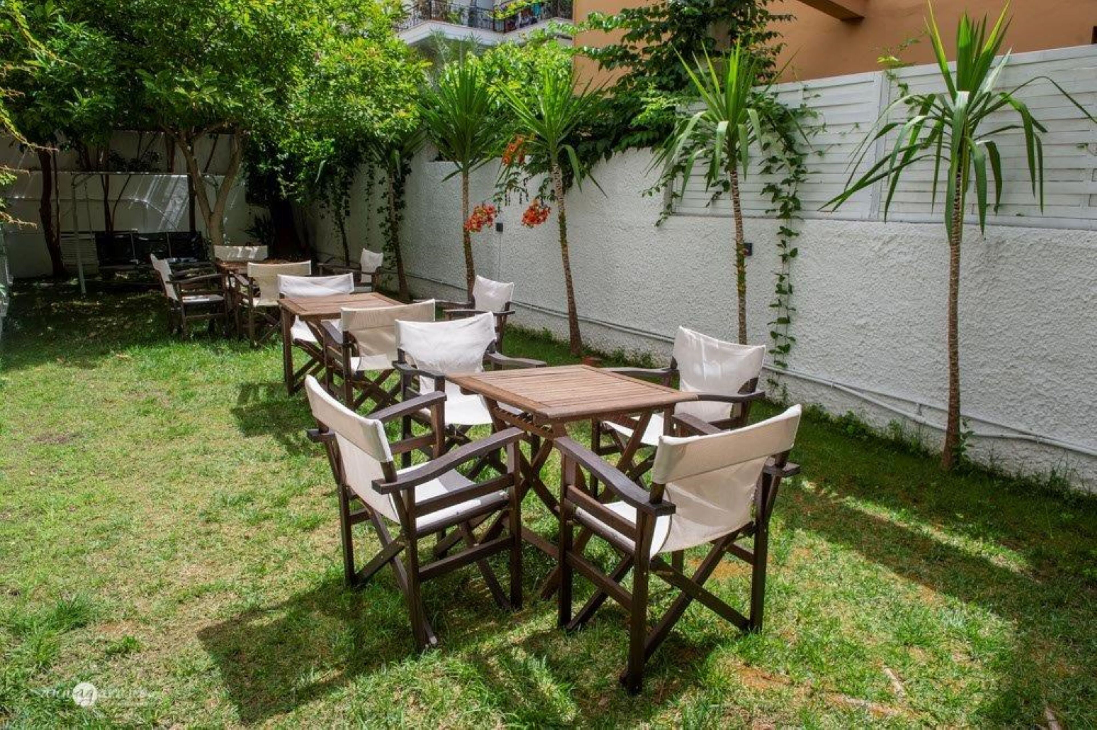 Eine Terrasse im Freien mit einem Holztisch und weißen Stühlen, umgeben von Pflanzen und einer weißen Mauer.