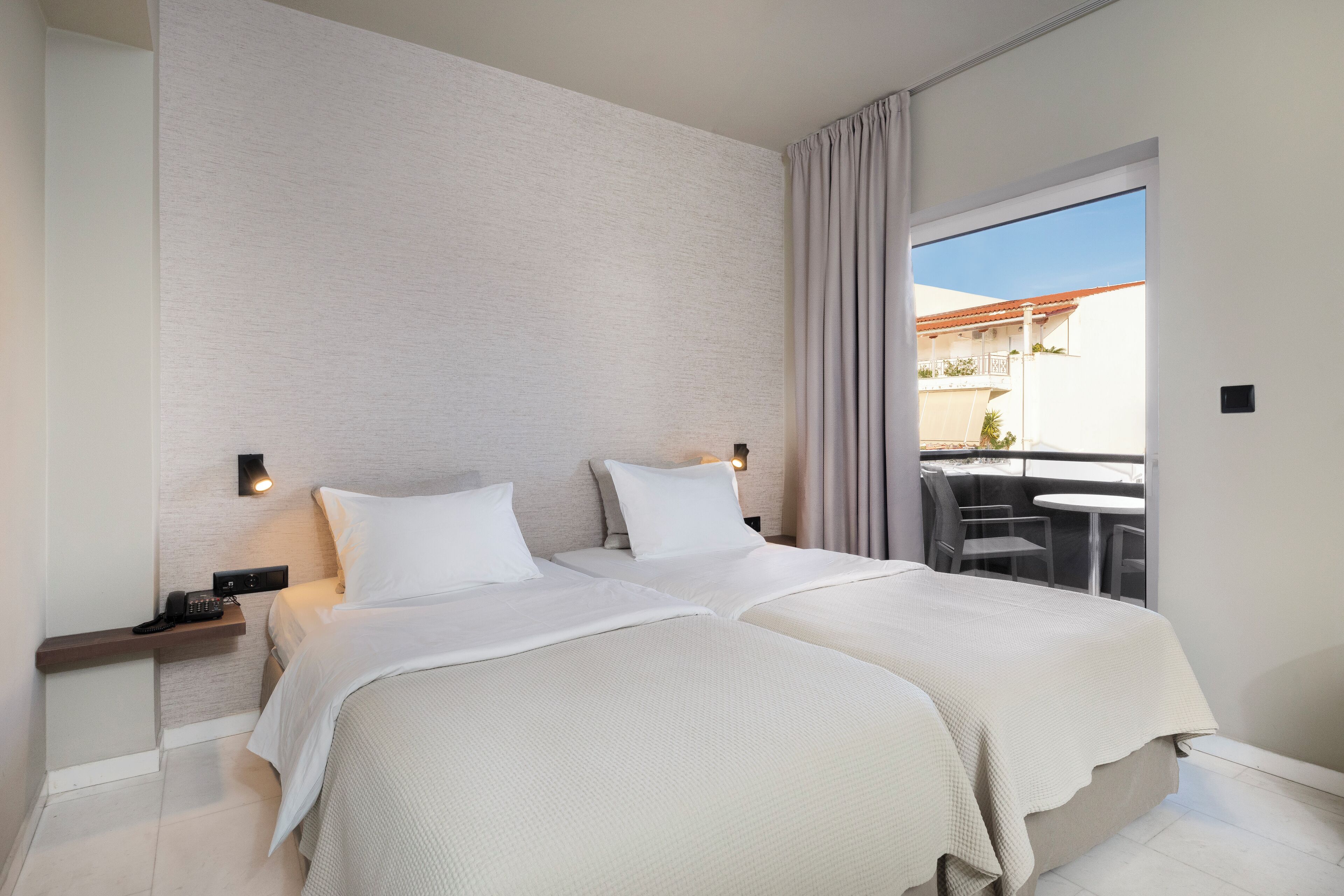 A hotel room with two beds, a small table, and a balcony with a view.