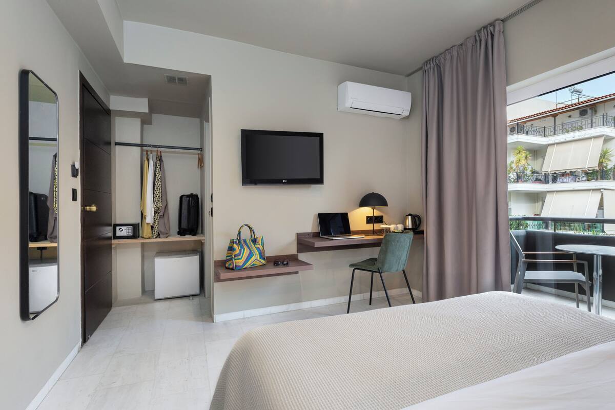 A hotel room with a bed, a desk with a laptop, a chair, a TV, and a balcony with a table and chairs.