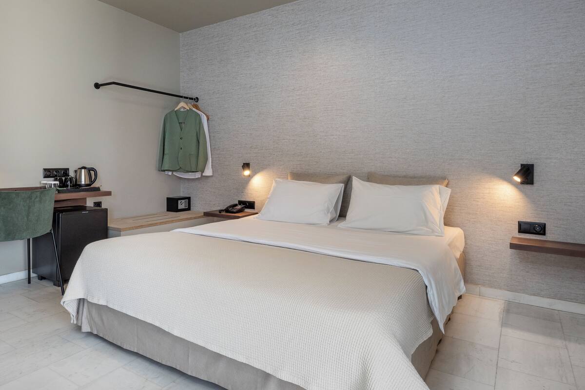 A modern hotel room with a large bed, bedside tables, a desk with a kettle, and a coat rack.